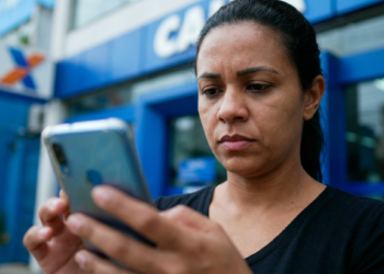 Mulher consulta o celular com expressão séria em frente à agência da Caixa