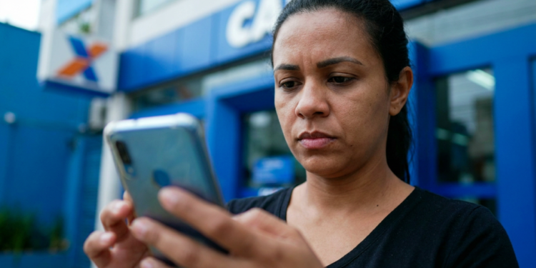 Mulher consulta o celular com expressão séria em frente à agência da Caixa