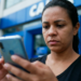 Mulher consulta o celular com expressão séria em frente à agência da Caixa
