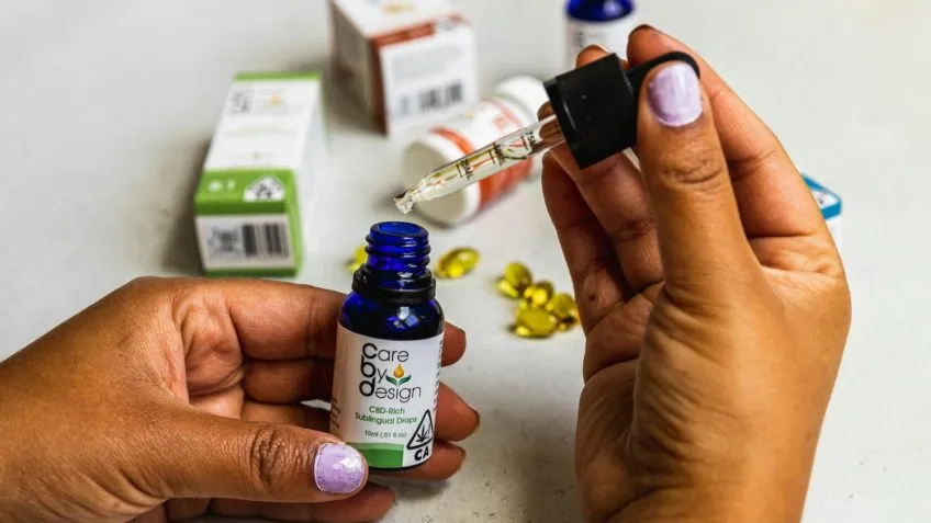 Mãos com unhas lilás segurando frasco azul de óleo CBD Care By Design e conta-gotas sobre mesa com cápsulas