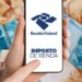 Mão segura um celular com a logomarca da Receita Federal