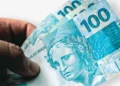 Close de uma mão segurando três cédulas de cem reais, destacando a Efígie da República. A imagem simboliza economia e pagamento de contas.