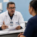 Perito médico do INSS de jaleco branco conversa com uma segurada à mesa durante atendimento de perícia