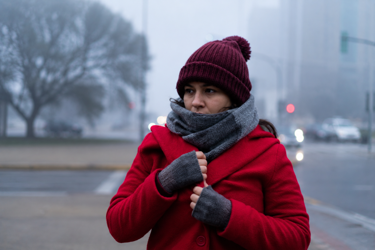 Mulher de casaco vermelho, gorro e cachecol se protege do frio em uma rua sob neblina