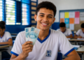 Estudante de uniforme sorri segurando duas cédulas de cem reais em uma sala de aula