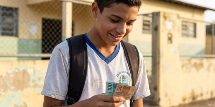 Estudante de uniforme sorri segurando notas de real e, ao redor, a fachada de uma escola