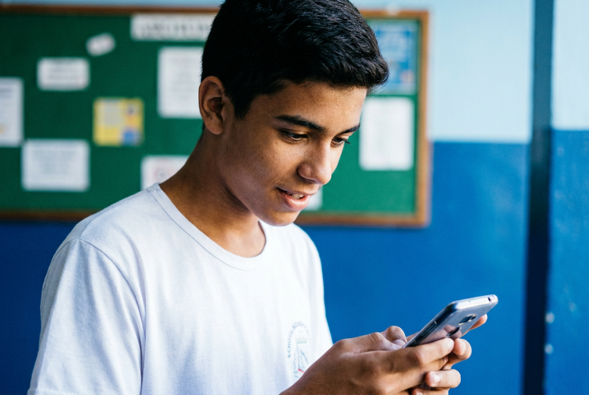 Estudante de uniforme usa o celular e, ao redor, um mural e a parede de uma sala de escola