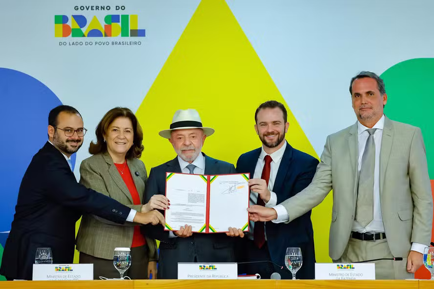 O Presidente da República, usando chapéu e terno, segura um documento oficial assinado junto a outros quatro representantes do governo. Ao fundo, um painel colorido com a logomarca "Governo do Brasil - Do lado do povo brasileiro".