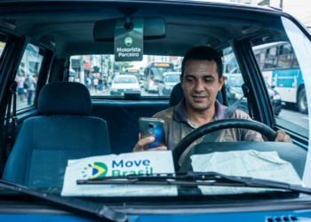 Motorista de aplicativo usa o celular dentro do carro, com cartaz do programa Move Brasil no painel.