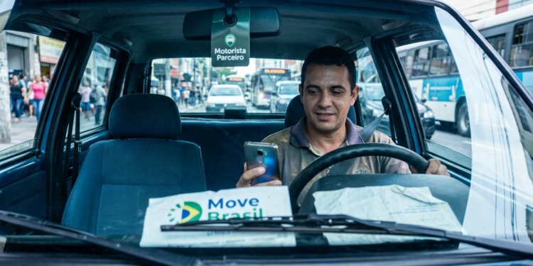 Motorista de aplicativo usa o celular dentro do carro, com cartaz do programa Move Brasil no painel.