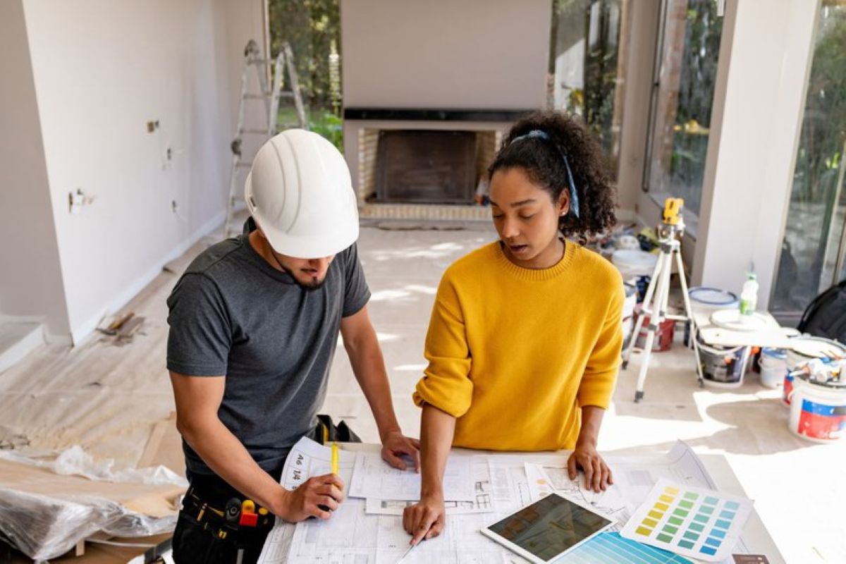 Homem com capacete e mulher analisando projeto de reforma dentro de uma residência em obras