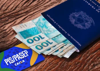 Carteira de Trabalho azul aberta com várias notas de 100 reais saindo de dentro. Ao lado, um cartão azul e amarelo com a inscrição "PIS/PASEP CAIXA". A imagem está sobre uma superfície de madeira.