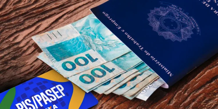 Carteira de Trabalho azul aberta com várias notas de 100 reais saindo de dentro. Ao lado, um cartão azul e amarelo com a inscrição "PIS/PASEP CAIXA". A imagem está sobre uma superfície de madeira.