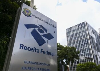 Placa da Receita Federal do Brasil diante de prédio administrativo e árvores em área urbana