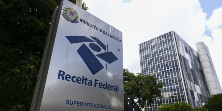 Placa da Receita Federal do Brasil diante de prédio administrativo e árvores em área urbana