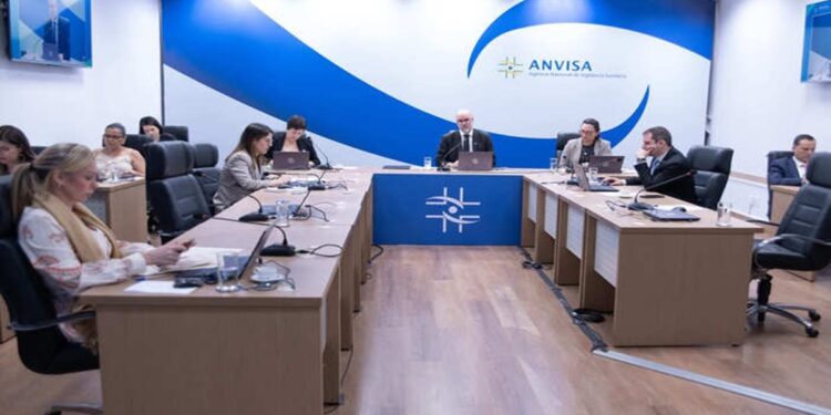 Diretoria da Anvisa reunida em sala de reuniões oficial com logomarca azul ao fundo e computadores sobre a mesa