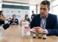 Homem exibe inalador e frascos rotulados como proibidos em mesa com microfones da Anvisa, coletiva ao fundo