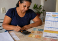 Mulher adulta usa calculadora ao lado de dinheiro em espécie e conta de energia elétrica detalhada sobre a mesa