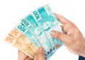 Mãos segurando cédulas de 50 e 100 reais em fundo branco.