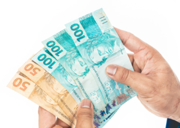 Mãos segurando cédulas de 50 e 100 reais em fundo branco.