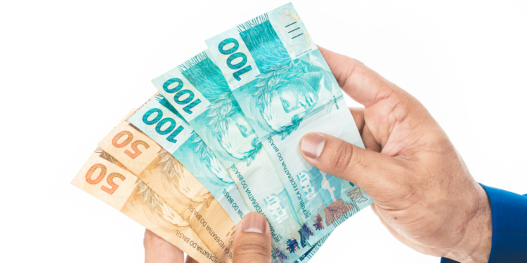 Mãos segurando cédulas de 50 e 100 reais em fundo branco.