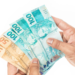 Mãos segurando cédulas de 50 e 100 reais em fundo branco.