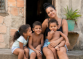 Mulher abraça três crianças sentadas na porta de casa simples, todos sorrindo