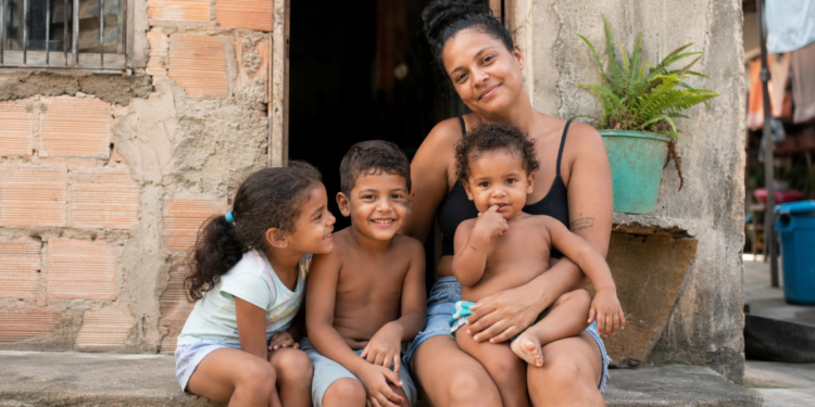 Mulher abraça três crianças sentadas na porta de casa simples, todos sorrindo