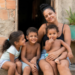Mulher abraça três crianças sentadas na porta de casa simples, todos sorrindo