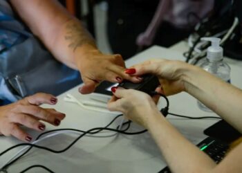 Mão de mulher sendo posicionada em leitor biométrico com manicure vermelha sobre mesa branca com computador