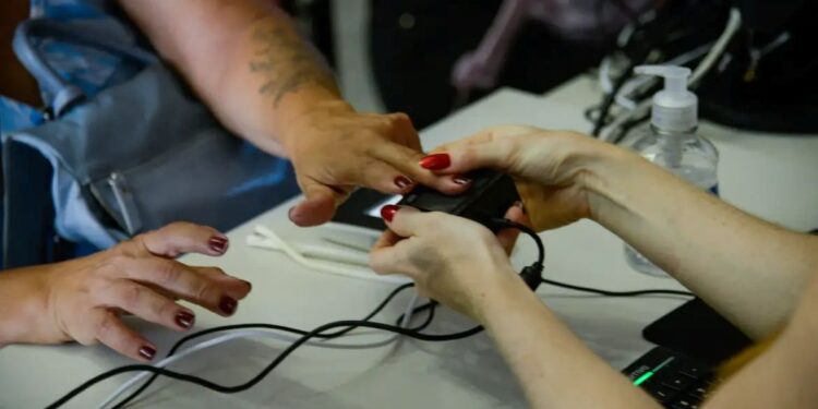 Mão de mulher sendo posicionada em leitor biométrico com manicure vermelha sobre mesa branca com computador