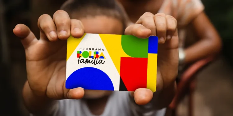 Mulher segurando cartão do Bolsa Família em destaque, ilustrando risco de bloqueio do benefício por pendências na área da saúde.