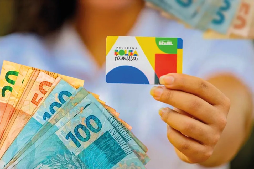 Pessoa segurando cartão do Bolsa Família ao lado de cédulas de real em foco