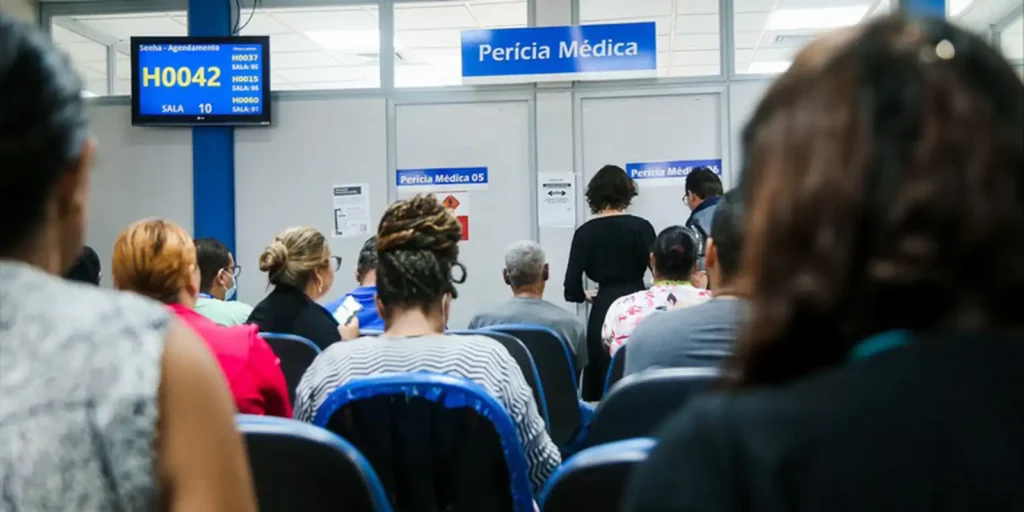 Pessoas de diferentes idades aguardam sentadas em frente à porta de perícia médica do INSS, com painel de senhas visível