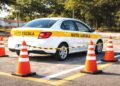 Veículo branco com faixa "AUTO ESCOLA" fazendo manobra cercado por seis cones laranja em pista aberta