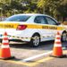 Veículo branco com faixa "AUTO ESCOLA" fazendo manobra cercado por seis cones laranja em pista aberta
