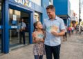 Homem e menina sorrindo, segurando notas de real, em frente à agência da Caixa Econômica Federal