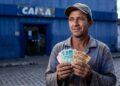 Trabalhador com boné e camisa azul segura notas de R$ 100 e R$ 50 em frente à agência da Caixa