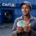 Trabalhador com boné e camisa azul segura notas de R$ 100 e R$ 50 em frente à agência da Caixa