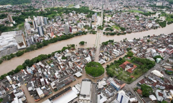 Vista aérea de cidade com rio marrom transbordado dividindo área urbana, ruas e prédios parcialmente submersos