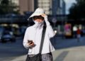 Mulher de roupa clara, chapéu grande e óculos escuros caminha olhando celular sob sol forte em avenida urbana