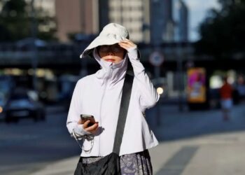 Mulher de roupa clara, chapéu grande e óculos escuros caminha olhando celular sob sol forte em avenida urbana