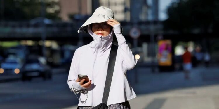 Mulher de roupa clara, chapéu grande e óculos escuros caminha olhando celular sob sol forte em avenida urbana