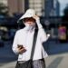 Mulher de roupa clara, chapéu grande e óculos escuros caminha olhando celular sob sol forte em avenida urbana