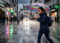 Mulher jovem atravessa a rua com guarda-chuva colorido durante tempestade de granizo, expressão tensa