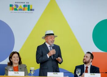 Presidente Lula, usando chapéu panamá, discursa ao microfone em evento do Governo do Brasil com painel colorido ao fundo