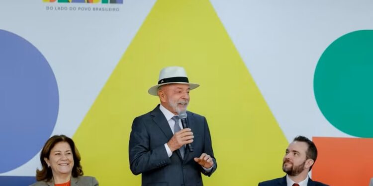 Presidente Lula, usando chapéu panamá, discursa ao microfone em evento do Governo do Brasil com painel colorido ao fundo