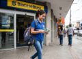 Jovem com uniforme escolar e mochila sorri segurando notas de dinheiro na frente do Banco do Brasil