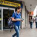 Jovem com uniforme escolar e mochila sorri segurando notas de dinheiro na frente do Banco do Brasil