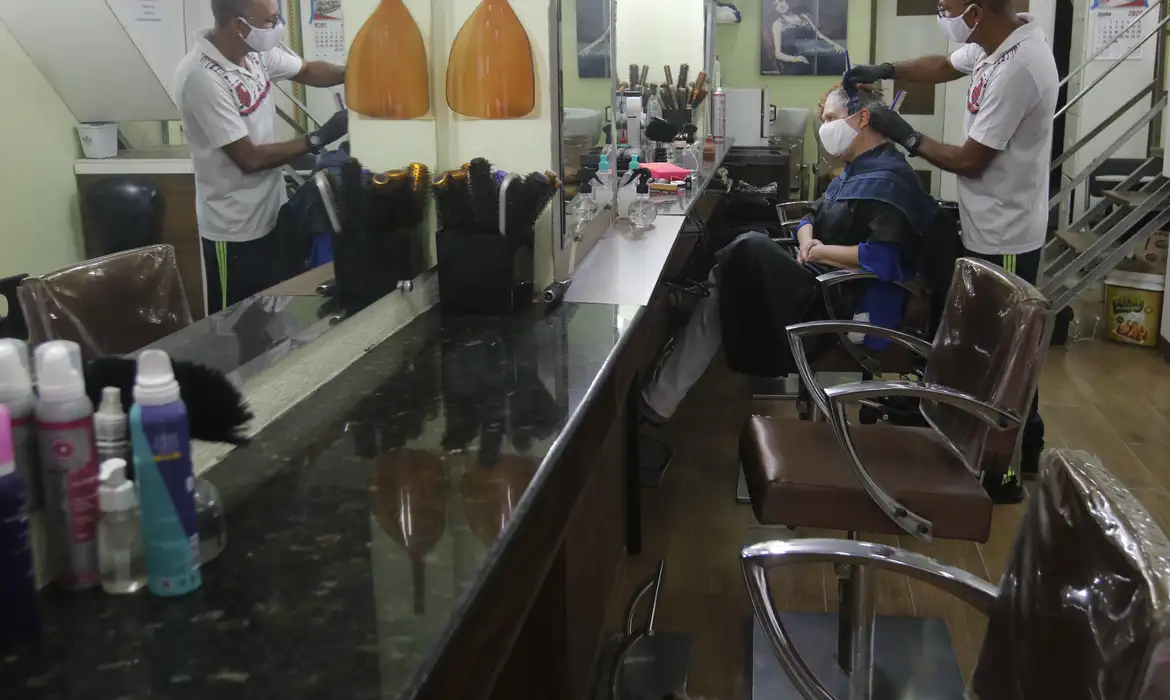 Cabeleireiro de máscara aplica produto no cabelo de mulher sentada em salão, ambos protegidos